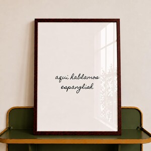 May include: A rectangular wall art print in a dark reddish-brown frame featuring the black cursive text "aqui hablamos espanglish" centered on a white background. The framed print is positioned on a dark olive green metal shelf with gold edges.