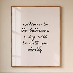 May include: A rectangular wall art print in a thin wooden frame featuring the black cursive text: welcome to the bathroom, a dog will be with you shortly. The print is displayed on a light-colored wall above a wooden stool and a white bathtub.