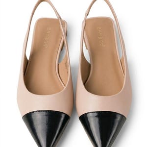 Puede incluir: Un par de zapatos planos de mujer con punta y correa trasera. Los zapatos son de dos tonos, con una parte superior rosa pálido y una puntera de charol negro. El nombre de la marca "Bamboo" está impreso en la plantilla.