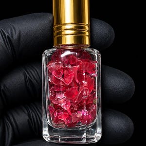 Puede incluir: Un frasco de vidrio transparente con aplicador de bola y tapa dorada brillante, relleno de pequeños fragmentos de piedras preciosas de color rojo oscuro translúcido. El frasco rectangular de esquinas redondeadas destaca sobre un fondo negro.