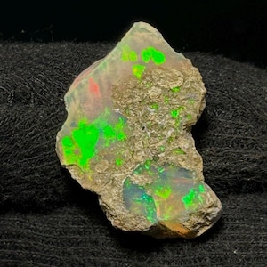 Könnte beinhalten: Ein rohes, unregelmäßiges Opal-Exemplar mit einer rauen, texturierten beigefarbenen Matrixoberfläche. Der Stein zeigt lebhafte, schillernde Blitze in Neongrün sowie subtile Nuancen von Orange und Rot auf seiner unebenen, teilweise polierten Fläche.