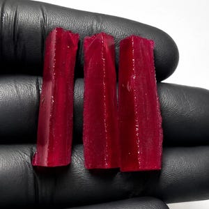 May include: Three elongated, raw, deep red gemstone crystals with a translucent, faceted appearance. Each crystal has a vertical, columnar structure with sharp, uneven edges and a glossy, reflective surface, displayed against a plain background.