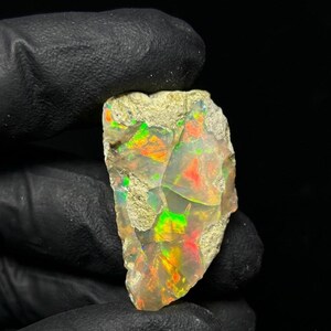 Könnte beinhalten: Ein roher, unpolierter Opal-Edelstein mit einer durchscheinenden, hellen Basis und leuchtenden Blitzen in Neongrün, Orange und Rot. Der Stein hat eine unregelmäßige, gezackte Form mit rauen Kanten und einer natürlichen, unebenen Oberfläche.