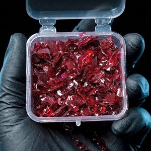 Puede incluir: Un pequeño recipiente cuadrado de plástico transparente lleno de numerosos fragmentos irregulares y dentados de vidrio rojo oscuro translúcido. El recipiente cuenta con una tapa abatible abierta que revela los fragmentos reflectantes de color rubí.