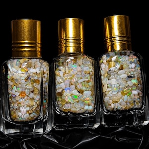 Könnte beinhalten: Drei durchsichtige Glasfläschchen mit Roll-on-Aufsatz und goldfarbenen Verschlüssen, gefüllt mit kleinen, polierten, irisierenden Opal-Edelsteinsplittern. Die Steine zeigen weiße, cremefarbene und durchscheinende Töne mit grünen, blauen und orangen Lichtreflexen.