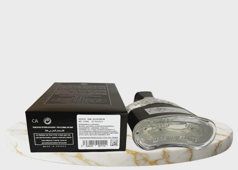 May include: A black rectangular perfume box with white text and a barcode next to a clear glass perfume bottle with a black cap. The box displays the text Aventus 100ml Eau de Parfum, ingredients, and Made in France. The bottle base is visible.