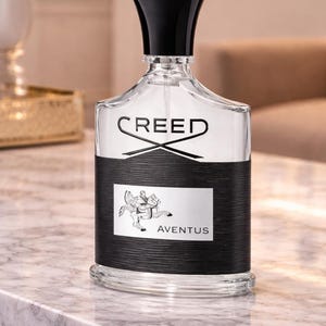 May include: A clear glass perfume bottle with a black cap and a black textured band around the center. The label features the word CREED at the top, an illustration of a person riding a horse in the middle, and the word AVENTUS at the bottom.