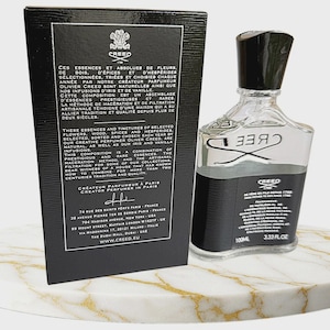 May include: A clear glass perfume bottle with a black cap and a black label featuring the Creed logo and 3.33 fl oz text, positioned next to a tall black rectangular box with white text detailing the brand history and locations in Paris.