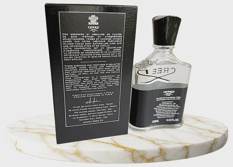 May include: A clear glass perfume bottle with a black cap and a black label featuring the Creed logo and 3.33 fl oz text, positioned next to a tall black rectangular box with white text detailing the brand history and locations in Paris.
