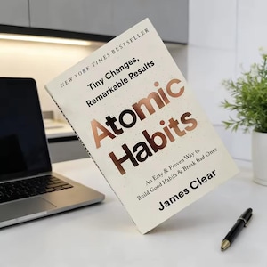 May include: A hardcover book titled "Atomic Habits" by James Clear, with the text "Tiny Changes, Remarkable Results." The book is open, displaying the title in copper-colored letters. A laptop and pen are also visible.