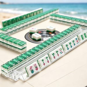 K&ouml;nnte beinhalten: Ein Mahjong-Set mit wei&szlig;en Spielsteinen und gr&uuml;nen, durchscheinenden R&uuml;ckseiten, die in Reihen auf einem beigen Tisch angeordnet sind. Die Steine zeigen verschiedene bunte Blumen- und Bambusmotive. Einige liegen in Kunststoffhaltern.