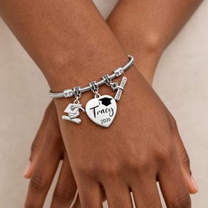 May include: Silver charm bracelet with graduation-themed charms. Charms include a graduation cap, a heart pendant with the name "Tracy" and the year "2026", and a small diploma. The bracelet is worn on a wrist.