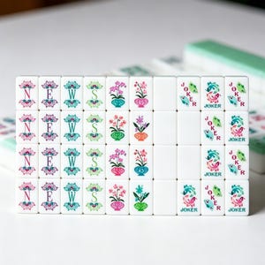 K&ouml;nnte beinhalten: Ein Set aus wei&szlig;en, rechteckigen Mahjong-Spielsteinen, die in einem Raster angeordnet sind. Die Steine zeigen bunte Druckmotive, darunter die Buchstaben N, E, W und S, Orchideen in T&ouml;pfen, leere Fl&auml;chen und ein Joker-Motiv mit stilisierten Fischen.