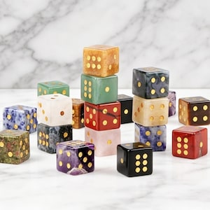 May include: A collection of colorful dice made from various materials, including marble and stone. Each die features gold-colored dots and comes in shades of green, red, black, and blue. The dice are arranged on a white marble surface.