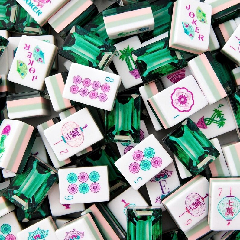 K&ouml;nnte beinhalten: Eine Sammlung von Mahjong-Spielsteinen, bestehend aus wei&szlig;en Steinen mit bunten Blumen-, Laternen- und Joker-Motiven sowie durchscheinenden smaragdgr&uuml;nen Steinen mit facettierter, edelsteinartiger Oberfl&auml;che und rechteckiger Form.