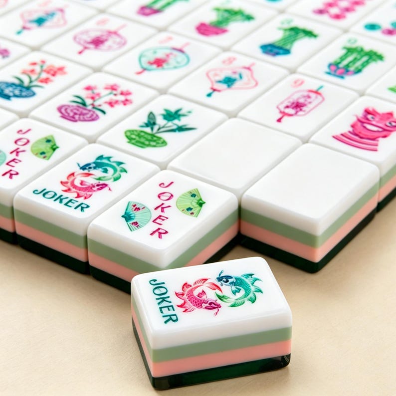 K&ouml;nnte beinhalten: Ein Set aus wei&szlig;en Mahjong-Spielsteinen mit geschichteten gr&uuml;nen, rosafarbenen und dunkelgr&uuml;nen Unterseiten. Die Steine zeigen farbige, eingravierte Illustrationen von Blumen, Laternen und Fischen. Einige Steine tragen das Wort JOKER in roter oder gr&uuml;ner Schrift.