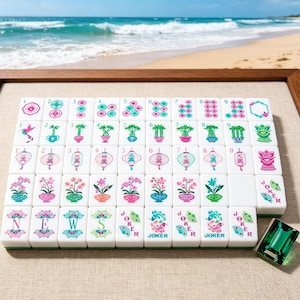 K&ouml;nnte beinhalten: Ein Set aus wei&szlig;en Mahjong-Spielsteinen in einem Raster, verziert mit bunten Illustrationen von Blumen, Bambus, Laternen und dem Wort JOKER. Ein einzelner, durchscheinender gr&uuml;ner Spielstein liegt separat auf einer strukturierten beigen Fl&auml;che.