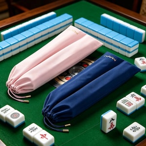 May include: A close-up shot of a Mahjong set on a green surface. The set includes blue and white tiles, along with pink and navy blue drawstring pouches. The pouches have gold-colored accents and the word "MAH" printed on them. The tiles are white with blue edges.