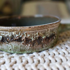 May include: A round ceramic bowl with a single handle, featuring a textured, mottled glaze in shades of sage green, brown, and tan. The exterior surface displays a prominent, rough, and craggy band of dark brown and green glaze around the middle.
