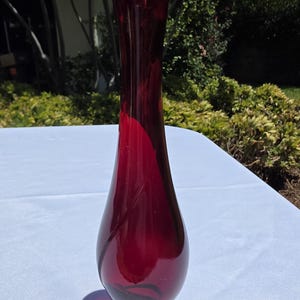Puede incluir: Un jarrón de cristal rojo rubí, alto y esbelto, con una base bulbosa que se estrecha hacia un cuello largo y estrecho con un borde acampanado e inclinado. El cristal transparente tiene un acabado liso y pulido con un patrón de remolinos.