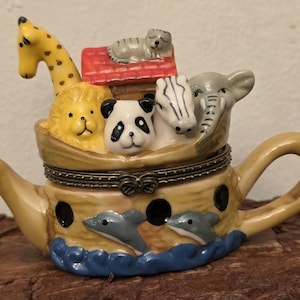 May include: A ceramic trinket box shaped like a teapot, featuring a boat filled with animal figurines. The animals include a giraffe, lion, panda, zebra, elephant, and a cat on a red roof. Dolphins swim on the base, with a beige and blue color scheme.