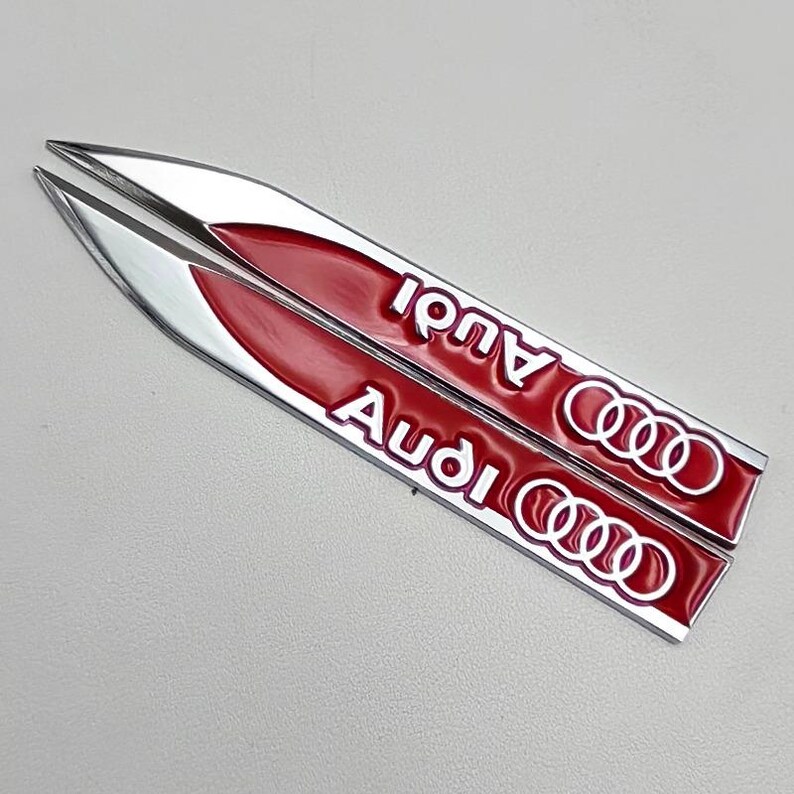 May include: Two silver and red Audi car emblems. The emblems are elongated and feature the Audi logo and the brand name in white lettering. The background is a light gray.