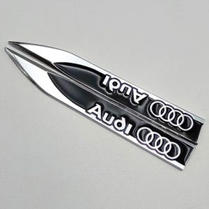 May include: Two silver and black Audi car emblems. Each emblem is a long, pointed shape with the word "Audi" in white lettering and the Audi logo. The emblems are on a light gray background.