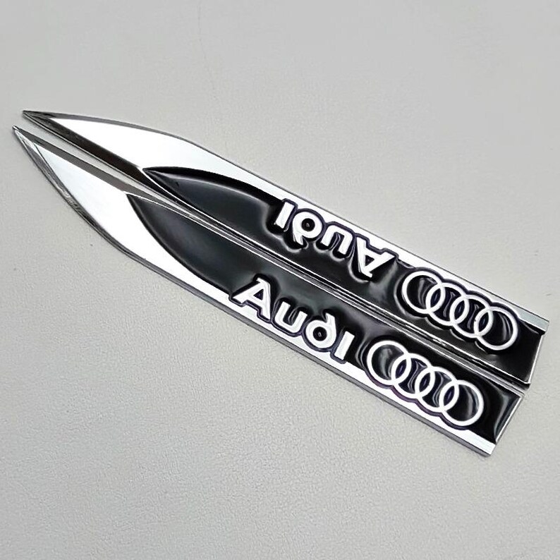 May include: Two silver and black Audi car emblems. Each emblem is a long, pointed shape with the word "Audi" in white lettering and the Audi logo. The emblems are on a light gray background.
