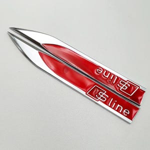 May include: Two silver and red S line car emblems. The emblems are elongated with a pointed end. The red section has the text "S line" in white. The emblems are likely for a car.