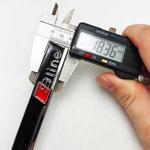 May include: A black and silver S line emblem with a red accent is measured with a digital caliper. The digital display shows a measurement of 18.36 mm. The emblem has the text "S line" in a stylized font.