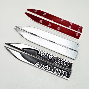 May include: A set of silver and black Audi car emblems. The black emblems have the Audi logo and text in white. Two silver emblems and two red adhesive strips are also visible.