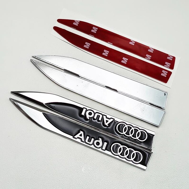 May include: A set of silver and black Audi car emblems. The black emblems have the Audi logo and text in white. Two silver emblems and two red adhesive strips are also visible.