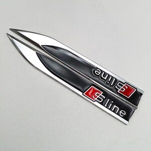 May include: Two silver and black S line car emblems. The emblems have a black background with the text "S line" in white and a red square with the "S" logo. The emblems are angled and have a pointed shape.