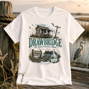 May include: White t-shirt featuring a detailed graphic of a rustic wooden structure with a pelican, railroad tracks, and the text "DRAWBRIDGE THE TRAIN STOPPED COMING Bay Area, California". The design includes a sign that reads "NO ROADS ONLY RAIL".