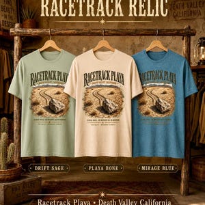 Puede incluir: Tres camisetas de manga corta en verde salvia, crema y azul, cada una con un gr&aacute;fico de una roca en un suelo des&eacute;rtico agrietado con el texto "RACETRACK PLAYA" y "DEATH VALLEY CALIFORNIA". Las camisetas est&aacute;n etiquetadas como "DRIFT SAGE", "PLAYA BONE" y "MIRAGE BLUE".