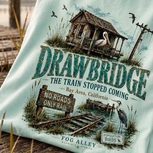 May include: Light blue t-shirt with a graphic design featuring a weathered wooden structure, a pelican, and the text "DRAWBRIDGE". The design includes the text "THE TRAIN STOPPED COMING" and "Bay Area, California".