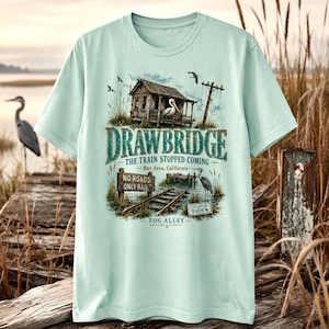May include: A light green t-shirt featuring a detailed graphic of a drawbridge scene. The design includes a weathered building, a pelican, and text that reads "DRAWBRIDGE" and "THE TRAIN STOPPED COMING." The shirt also has text that reads "Bay Area, California" and "NO ROADS ONLY RAIL."