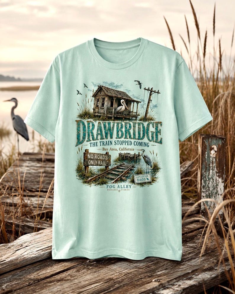 May include: A light green t-shirt featuring a detailed graphic of a drawbridge scene. The design includes a weathered building, a pelican, and text that reads "DRAWBRIDGE" and "THE TRAIN STOPPED COMING." The shirt also has text that reads "Bay Area, California" and "NO ROADS ONLY RAIL."