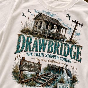 May include: White t-shirt featuring a detailed graphic of a rustic waterside scene. The design includes a weathered wooden house, a pelican, a heron, and the text "DRAWBRIDGE" and "THE TRAIN STOPPED COMING". Additional text reads "NO ROADS ONLY RAIL" and "POPULATION BIRDS".