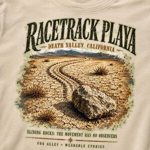 Puede incluir: Una camiseta beige con un gr&aacute;fico de Racetrack Playa en el Valle de la Muerte, California. El estampado muestra una roca sobre tierra agrietada con un rastro, y el texto "RACETRACK PLAYA", "SLIDING ROCKS: THE MOVEMENT HAS NO OBSERVERS".