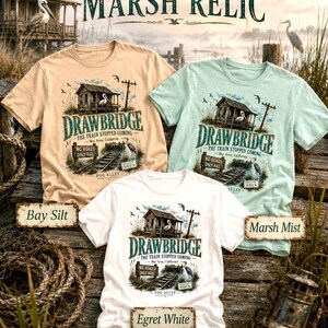 May include: Three t-shirts in beige, mint green, and white, each featuring a graphic of a weathered building, a pelican, and the text "DRAWBRIDGE". The text "THE TRAIN STOPPED COMING" and "NO ROADS ONLY RAIL" are also visible. The shirts are labeled "Bay Silt", "Marsh Mist", and "Egret White".