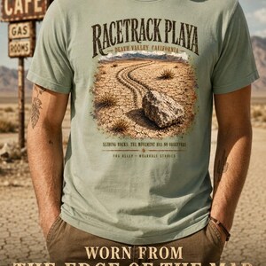Puede incluir: Una camiseta de manga corta color verde salvia con un gr&aacute;fico de una roca sobre un suelo des&eacute;rtico agrietado, con el texto RACETRACK PLAYA DEATH VALLEY CALIFORNIA, SLIDING ROCKS: THE MOVEMENT HAS NO OBSERVERS y FOG ALLEY WEARABLE STORIES impreso.