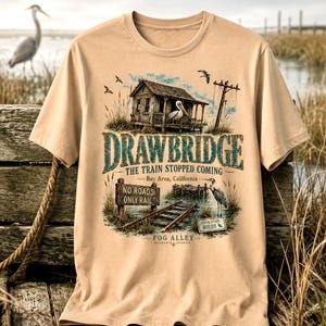 May include: Tan t-shirt featuring a detailed graphic of a weathered wooden structure with a pelican, railroad tracks, and the text "DRAWBRIDGE THE TRAIN STOPPED COMING Bay Area, California." Additional text includes "NO ROADS ONLY RAIL" and "FOG ALLEY WEARABLE STORIES."
