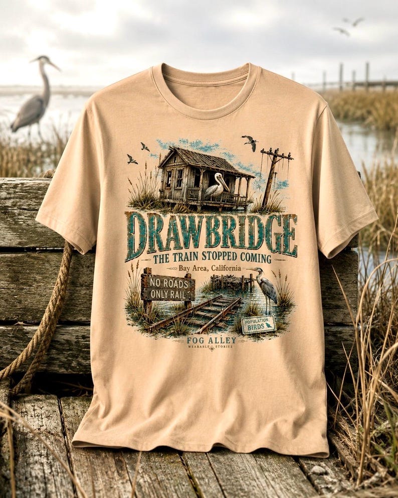 May include: Tan t-shirt featuring a detailed graphic of a weathered wooden structure with a pelican, railroad tracks, and the text "DRAWBRIDGE THE TRAIN STOPPED COMING Bay Area, California." Additional text includes "NO ROADS ONLY RAIL" and "FOG ALLEY WEARABLE STORIES."