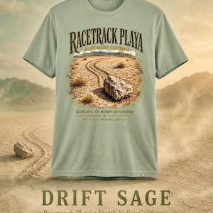 Puede incluir: Una camiseta de color verde salvia con un gr&aacute;fico de Racetrack Playa en el Valle de la Muerte, California. El dise&ntilde;o muestra un paisaje des&eacute;rtico con una roca deslizante y huellas, con el texto "RACETRACK PLAYA", "DEATH VALLEY CALIFORNIA" y "SLIDING ROCKS: THE MOVEMENT HAS NO OBSERVERS".