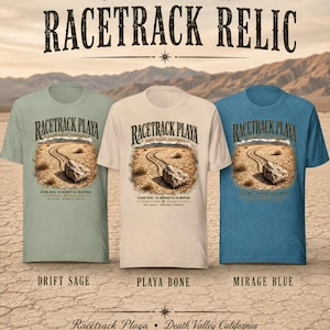 Puede incluir: Tres camisetas de manga corta en color verde salvia, hueso y azul espejismo. Cada camiseta presenta un gr&aacute;fico de una roca sobre un suelo des&eacute;rtico agrietado con el texto Racetrack Playa Death Valley California. El texto Choose Your Racetrack Relic aparece arriba.