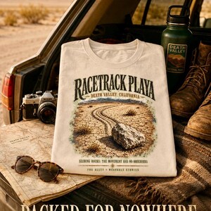 Puede incluir: Una camiseta de color crema con un gr&aacute;fico de Racetrack Playa en el Valle de la Muerte, California, que muestra una roca y un rastro, junto al texto "Racetrack Playa", "Sliding Rocks: The Movement Has No Observers" y "Fog Alley - Wearable Stories".