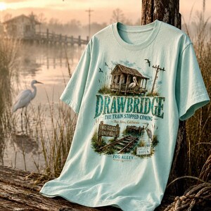 May include: A light blue t-shirt featuring a graphic of a rustic shack, a heron, and the text "DRAWBRIDGE THE TRAIN STOPPED COMING Bay Area, California". The design includes a railroad track and the words "NO ROADS ONLY RAIL".