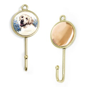 Op de afbeelding: Twee decoratieve wandhaken. Eén haak heeft een cirkelvormig ontwerp met een lachende gele Labrador Retriever, blauwe bloemen en het woord "Happy". De andere haak heeft een koperkleurig cirkelvormig ontwerp. Beide haken hebben een messing afwerking en een haakvorm.