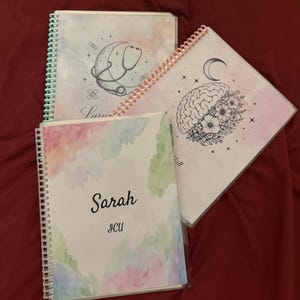 May include: Three spiral-bound notebooks with watercolour-style covers. One features a crescent moon and stethoscope, another a brain with flowers and a moon, and the third displays the name Sarah and the text ICU in black lettering.
